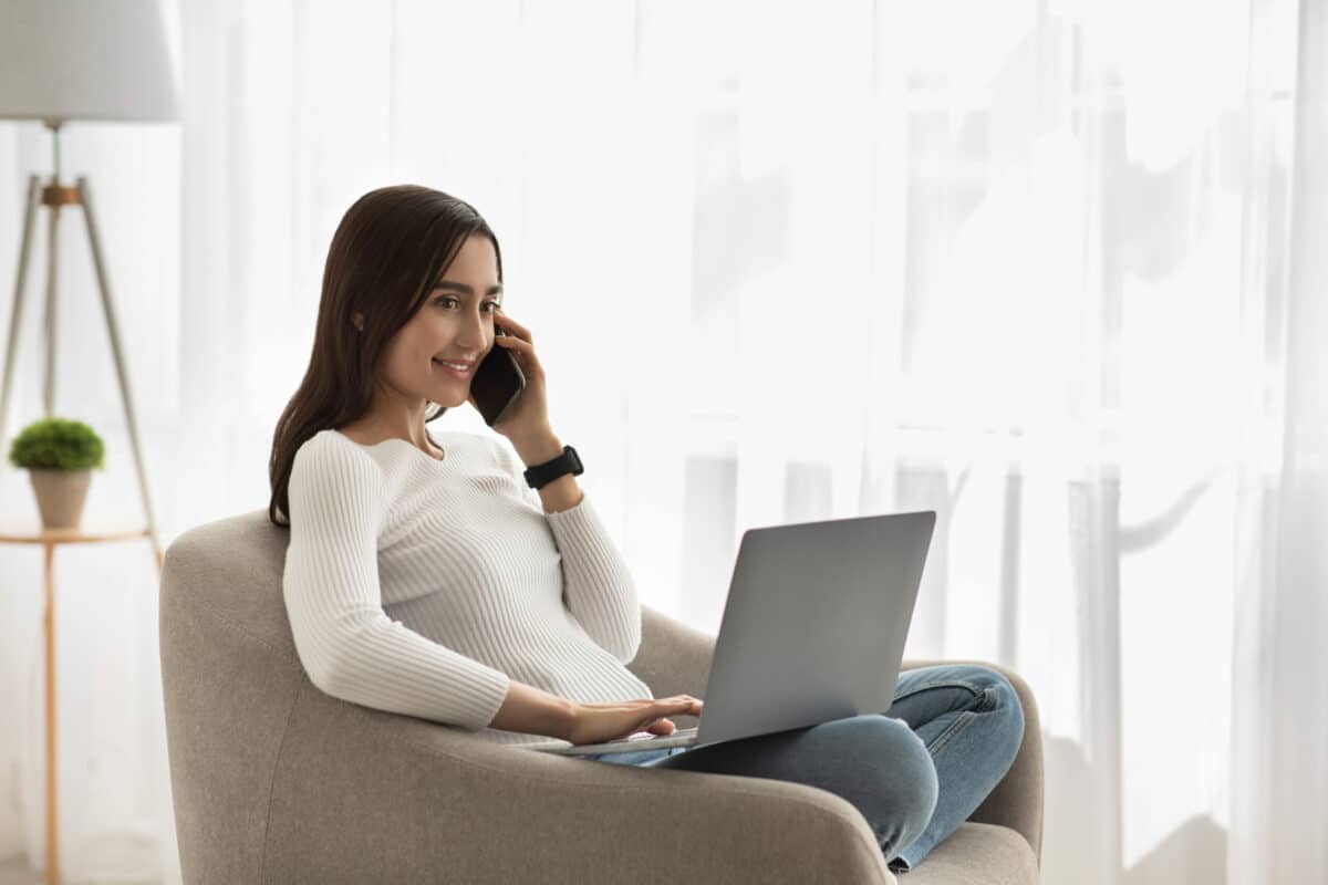 a woman, living alone at home sits on a chair on the phone with her laptop